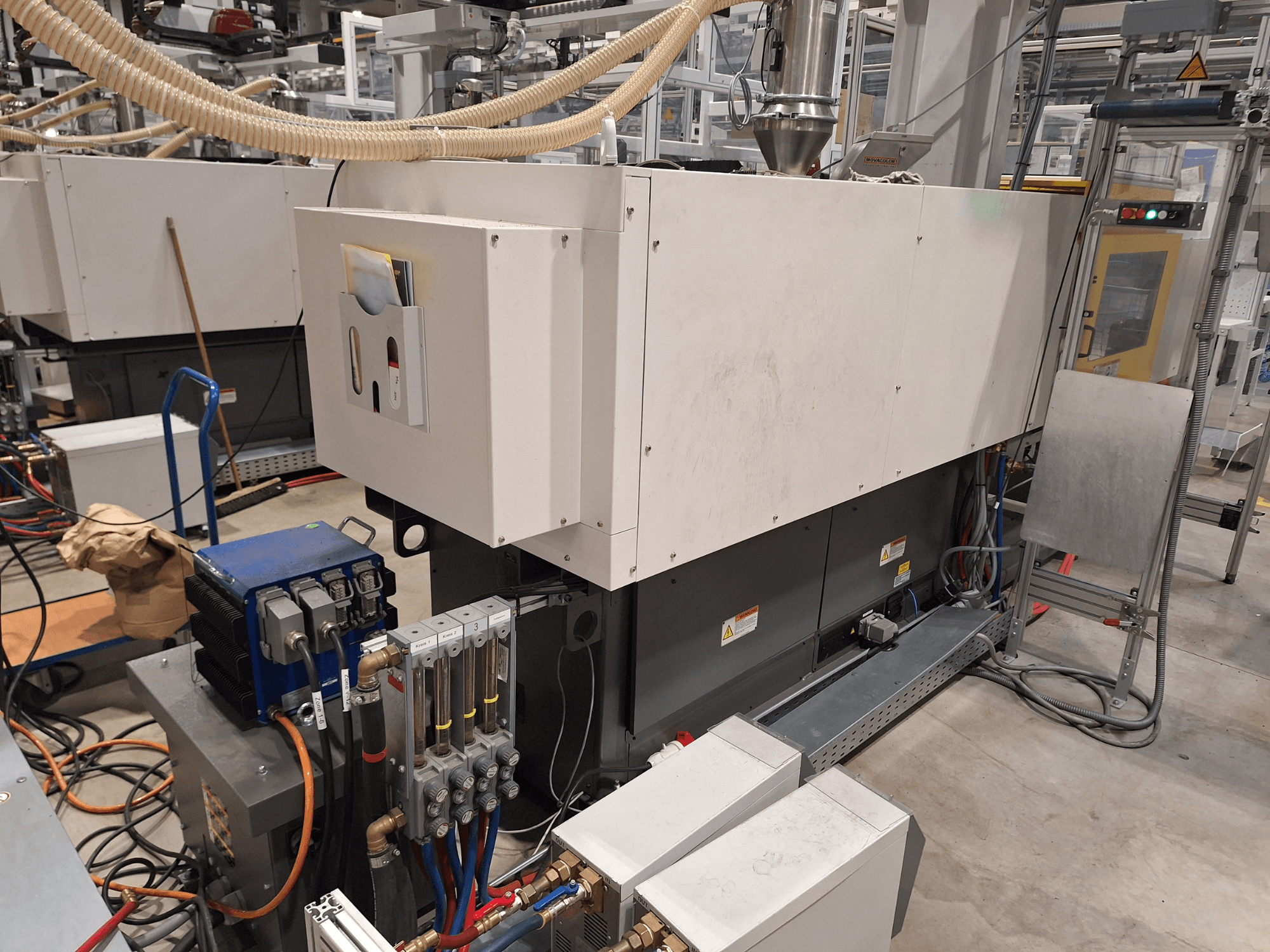 Industrial equipment in a factory setting; includes a large machine with control panel, hoses, and electrical connections, side view.